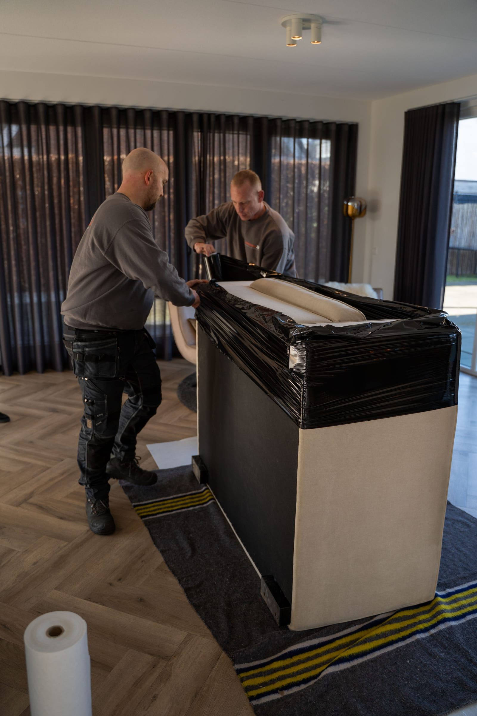 Twee verhuizers dragen een in folie verpakt meubel door de woonkamer