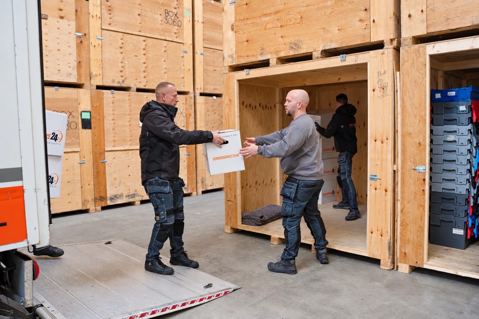 Verhuizers geven verhuisdozen door van de laadklep naar een houten opslagbox in de loods van Verhuisservice24